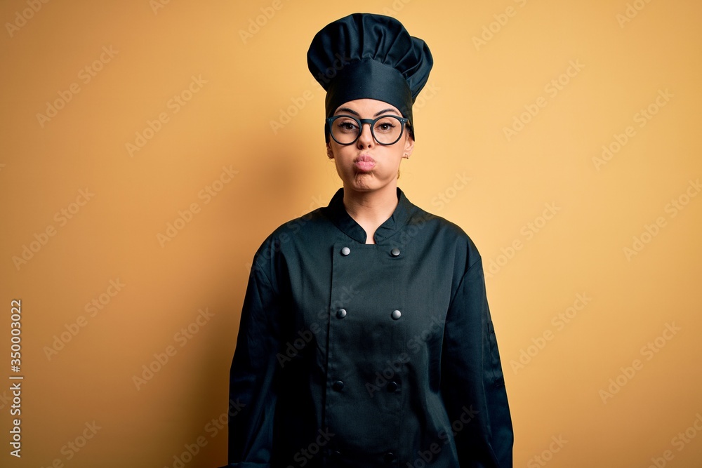 Young beautiful brunette chef woman wearing cooker uniform and hat over yellow background puffing cheeks with funny face. Mouth inflated with air, crazy expression. Young beautiful brunette chef woman wearing cooker uniform and hat over yellow background puffing cheeks with funny face. Mouth inflated with air, crazy expression.