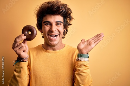 Young handsome man holding chocolate donut standing over isolated yellow background very happy and excited, winner expression celebrating victory screaming with big smile and raised hands