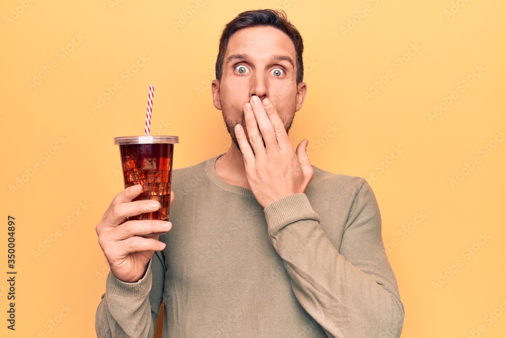 Young handsome man drinking cola refreshment beverage over isolated yellow background covering mouth with hand, shocked and afraid for mistake. Surprised expression Young handsome man drinking cola refreshment beverage over isolated yellow background covering mouth with hand, shocked and afraid for mistake. Surprised expression