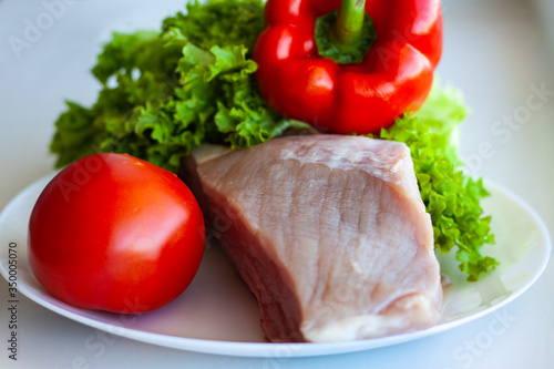 A piece of raw pork on a white plate with lettuce, tomato and sweet pepper. Healthy eating and cooking at home concept