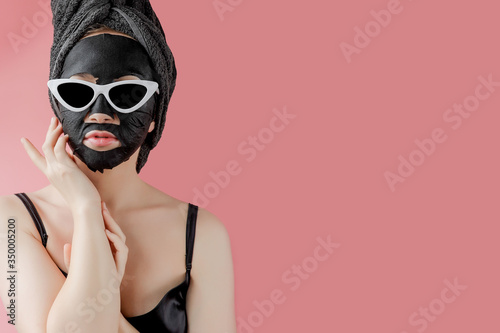 Young woman in glasses appling black cosmetic fabric facial mask on pink background. Face peeling mask with charcoal, spa beauty treatment, skincare, cosmetology. Close up