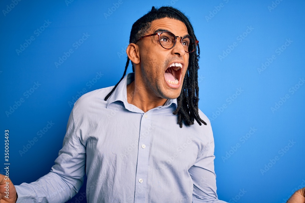 Young handsome african american man with dreadlocks wearing casual shirt and glasses crazy and mad shouting and yelling with aggressive expression and arms raised. Frustration concept. Young handsome african american man with dreadlocks wearing casual shirt and glasses crazy and mad shouting and yelling with aggressive expression and arms raised. Frustration concept.
