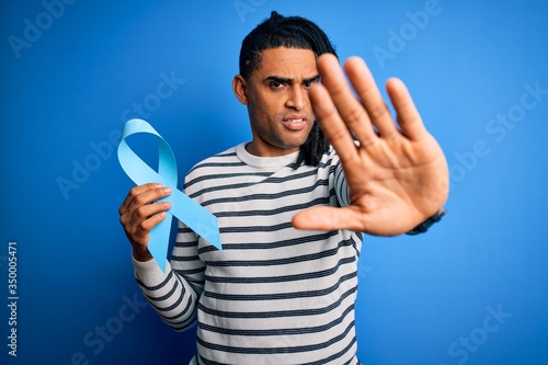 Young african american afro man with dreadlocks holding blue cancer ribbon with open hand doing stop sign with serious and confident expression, defense gesture