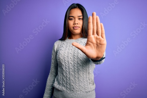 Young beautiful asian girl wearing casual sweater standing over isolated purple background doing stop sing with palm of the hand. Warning expression with negative and serious gesture on the face.