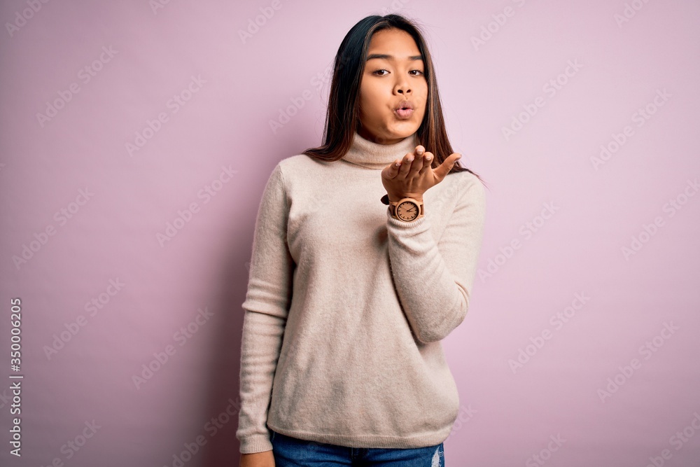 Young beautiful asian girl wearing casual turtleneck sweater over isolated pink background looking at the camera blowing a kiss with hand on air being lovely and sexy. Love expression. Young beautiful asian girl wearing casual turtleneck sweater over isolated pink background looking at the camera blowing a kiss with hand on air being lovely and sexy. Love expression.