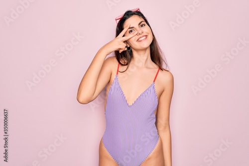 Young beautiful fashion girl wearing swimwear swimsuit and sunglasses over pink background Doing peace symbol with fingers over face, smiling cheerful showing victory