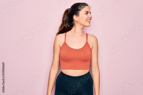 Young beautiful fitness woman wearing sport excersie clothes over pink background looking away to side with smile on face  natural expression. Laughing confident.