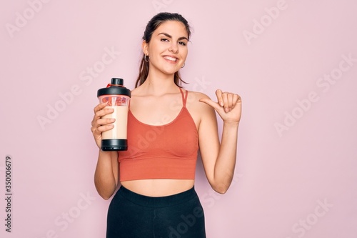 Young beautiful fitness woman wearing sport excersie clothes drinking protein shake with surprise face pointing finger to himself