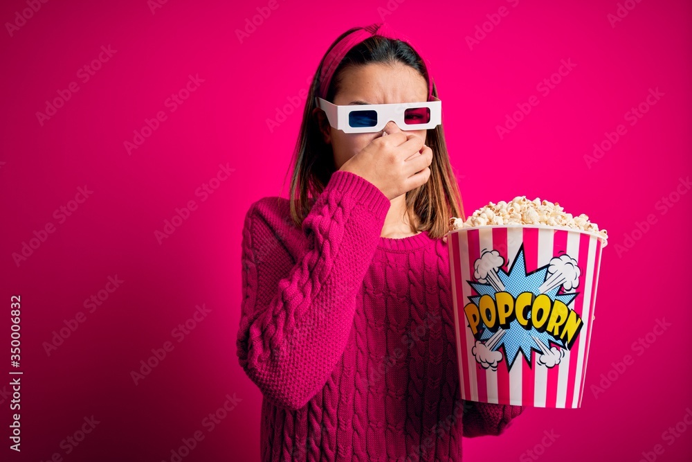 Obraz premium Young beautiful girl watching movie using 3d glasses eating box with popcorns smelling something stinky and disgusting, intolerable smell, holding breath with fingers on nose. Bad smell