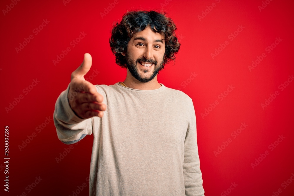 Young handsome man with beard wearing casual sweater standing over red background smiling friendly offering handshake as greeting and welcoming. Successful business. Young handsome man with beard wearing casual sweater standing over red background smiling friendly offering handshake as greeting and welcoming. Successful business.