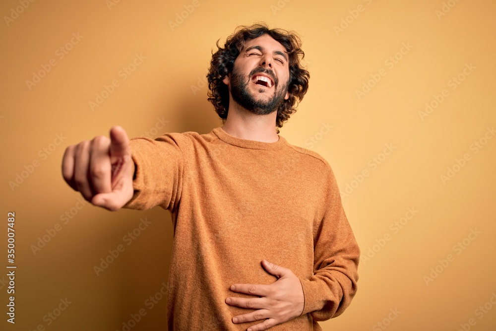 Young handsome man with beard wearing casual sweater standing over yellow background laughing at you, pointing finger to the camera with hand over body, shame expression Young handsome man with beard wearing casual sweater standing over yellow background laughing at you, pointing finger to the camera with hand over body, shame expression