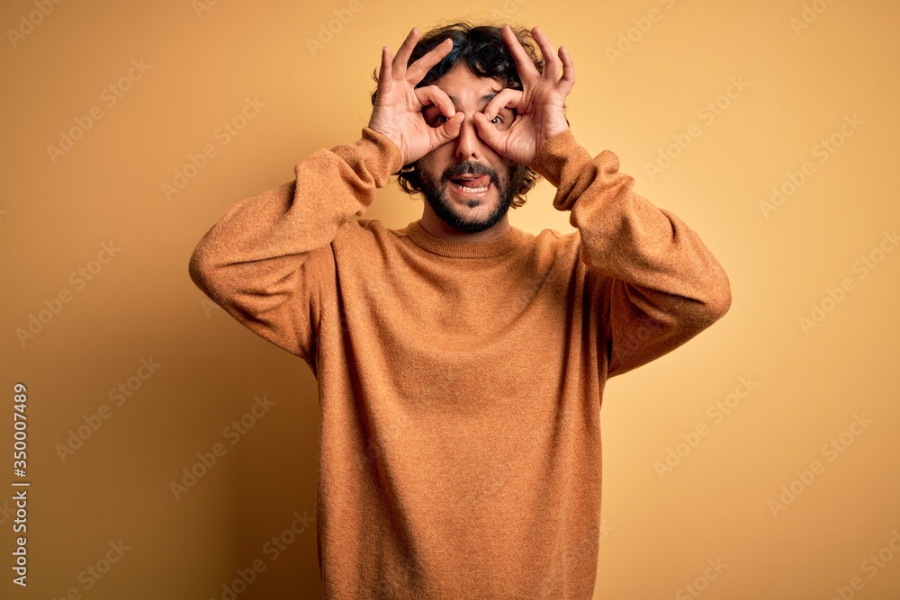 Young handsome man with beard wearing casual sweater standing over yellow background doing ok gesture like binoculars sticking tongue out, eyes looking through fingers. Crazy expression. Young handsome man with beard wearing casual sweater standing over yellow background doing ok gesture like binoculars sticking tongue out, eyes looking through fingers. Crazy expression.