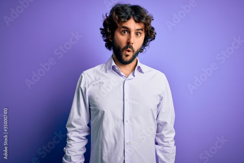 Young handsome business man with beard wearing shirt standing over purple background In shock face, looking skeptical and sarcastic, surprised with open mouth