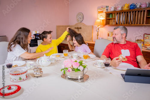 Happy family having breakfast together at home in the kitchen - Mother, son, father and daughter having a typical italian breakfast - Father is working wit  his tablet - The children pull their hair