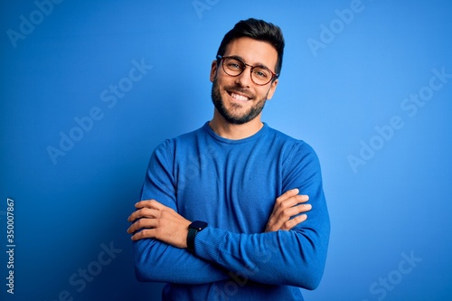 Bilde på lerret Young handsome man with beard wearing casual sweater and glasses over blue background happy face smiling with crossed arms looking at the camera