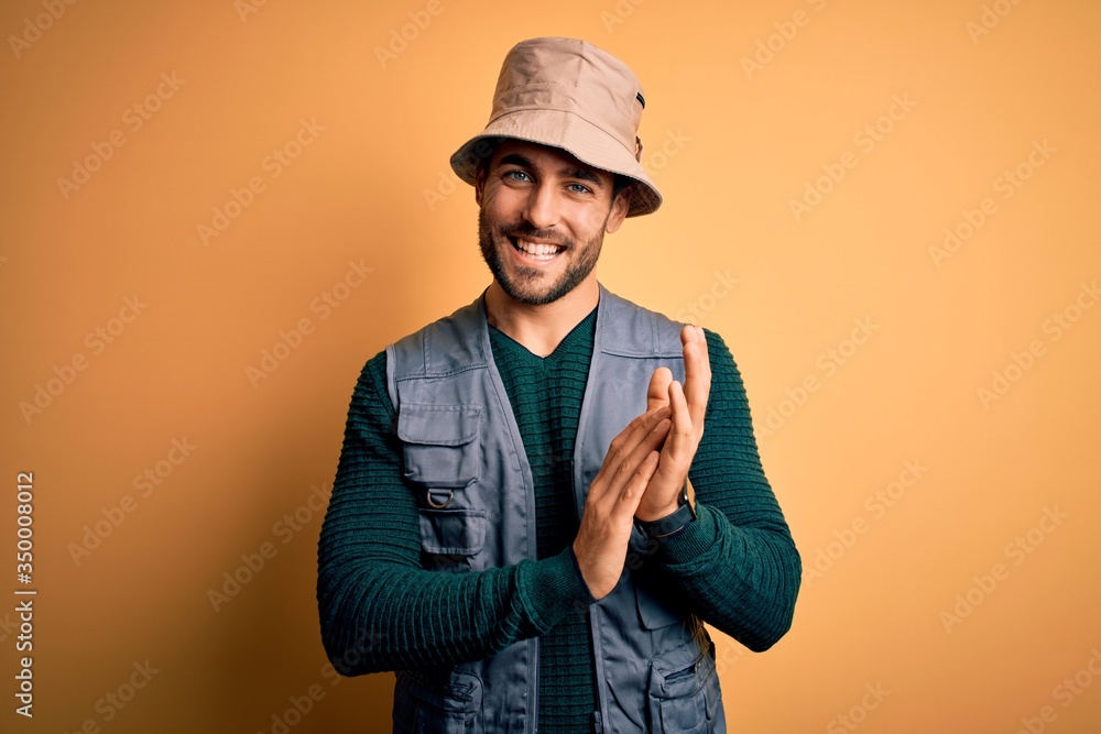 Handsome tourist man with beard on vacation wearing explorer hat over yellow background clapping and applauding happy and joyful, smiling proud hands together Handsome tourist man with beard on vacation wearing explorer hat over yellow background clapping and applauding happy and joyful, smiling proud hands together