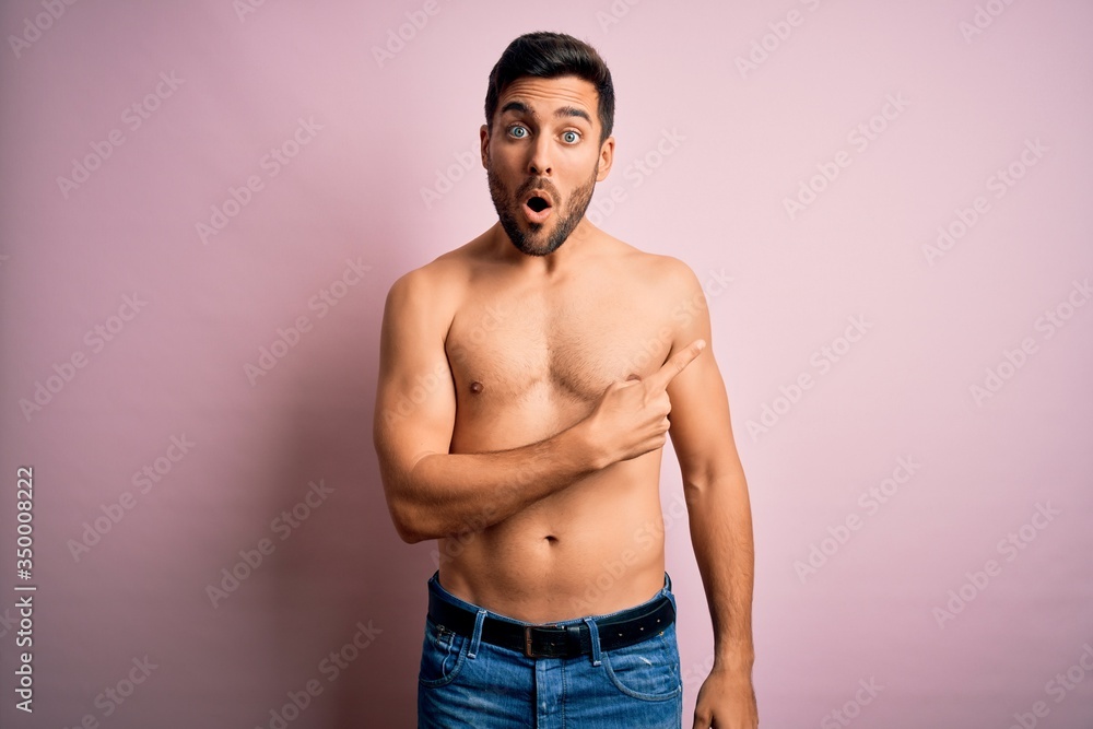 Young handsome strong man with beard shirtless standing over isolated pink background Surprised pointing with finger to the side, open mouth amazed expression.