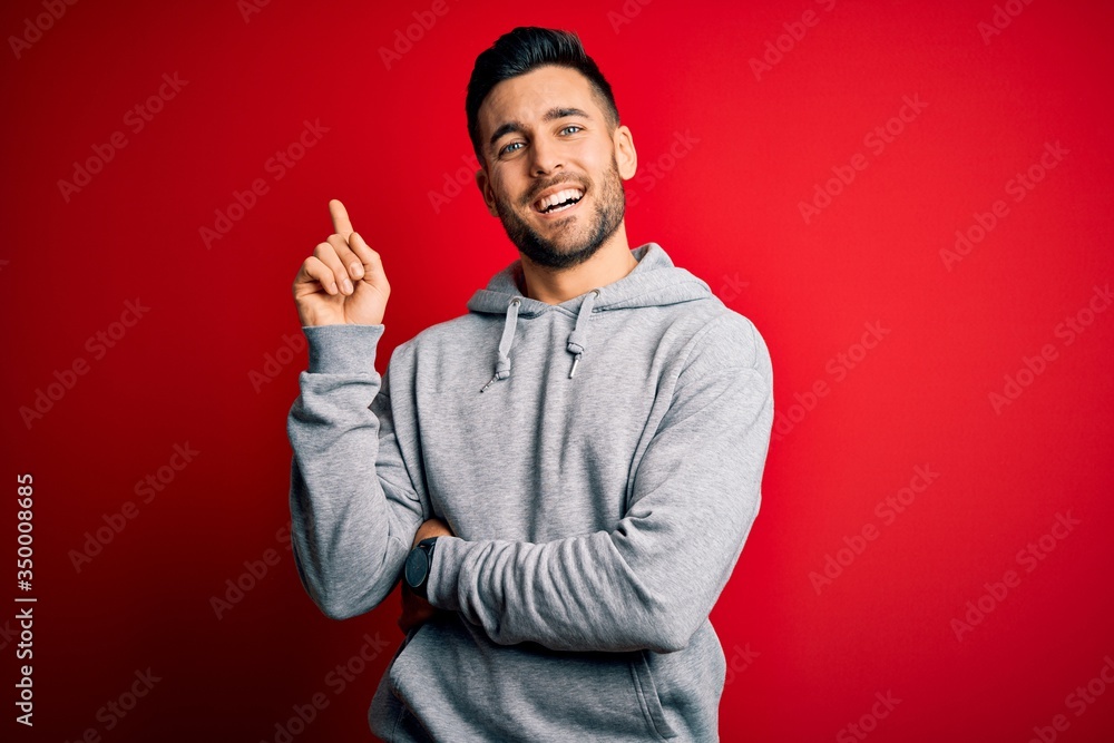 Young handsome sportsman wearing sweatshirt standing over isolated red background with a big smile on face, pointing with hand and finger to the side looking at the camera. Young handsome sportsman wearing sweatshirt standing over isolated red background with a big smile on face, pointing with hand and finger to the side looking at the camera.