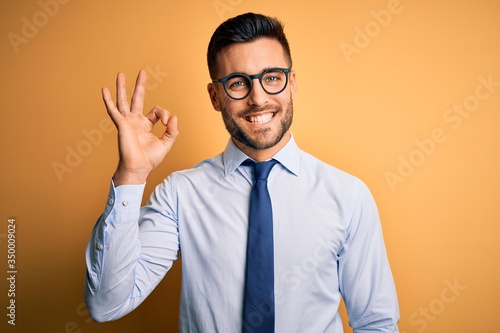 Young handsome businessman wearing tie and glasses standing over yellow background smiling positive doing ok sign with hand and fingers. Successful expression.