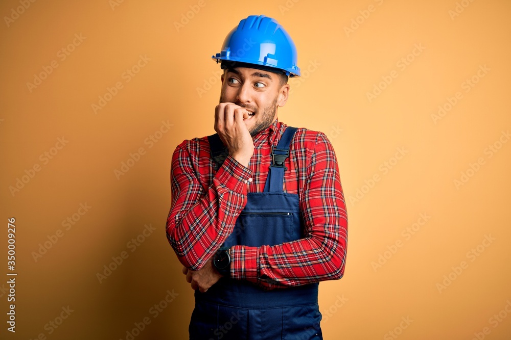 Young builder man wearing construction uniform and safety helmet over yellow background looking stressed and nervous with hands on mouth biting nails. Anxiety problem.