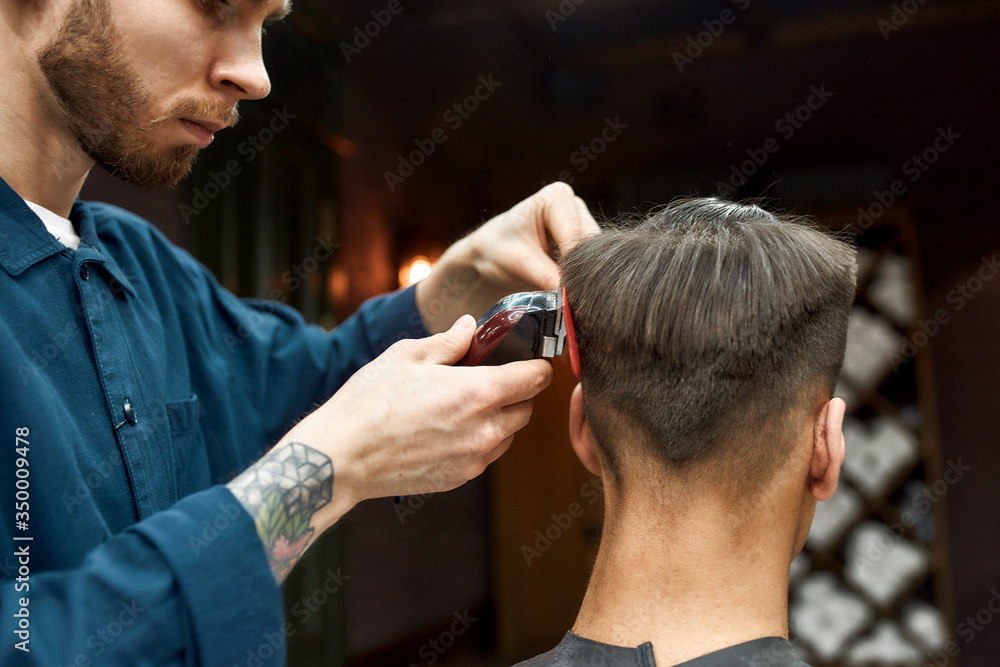 Master. Bearded barber with tattoo on his hand cutting clients hair with hair clipper. Barbershop. Back view Master. Bearded barber with tattoo on his hand cutting clients hair with hair clipper. Barbershop. Back view