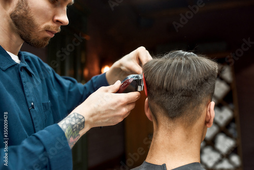 Master. Bearded barber with tattoo on his hand cutting clients hair with hair clipper. Barbershop. Back view