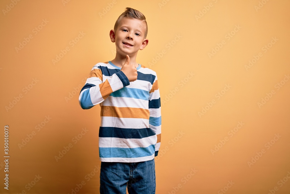 Young little caucasian kid with blue eyes wearing colorful striped shirt over yellow background doing happy thumbs up gesture with hand. Approving expression looking at the camera showing success. Young little caucasian kid with blue eyes wearing colorful striped shirt over yellow background doing happy thumbs up gesture with hand. Approving expression looking at the camera showing success.