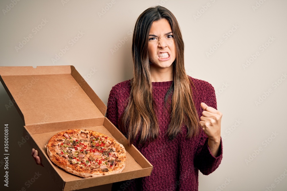 Young beautiful girl holding delivery box with Italian pizza standing over white background annoyed and frustrated shouting with anger, crazy and yelling with raised hand, anger concept Young beautiful girl holding delivery box with Italian pizza standing over white background annoyed and frustrated shouting with anger, crazy and yelling with raised hand, anger concept