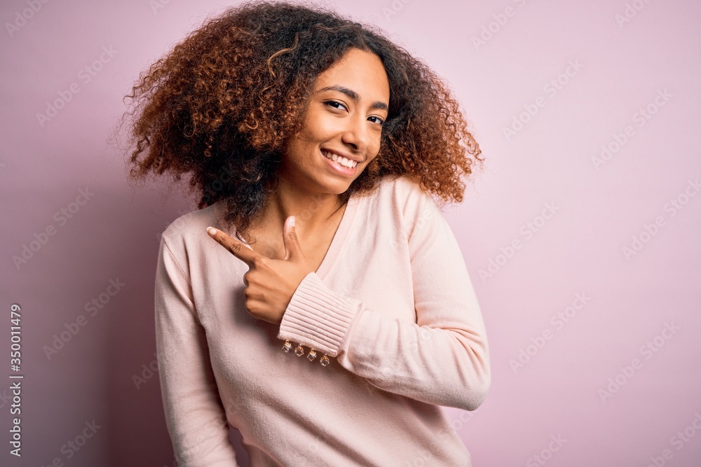 Young african american woman with afro hair wearing casual sweater over pink background cheerful with a smile of face pointing with hand and finger up to the side with happy and natural expression Young african american woman with afro hair wearing casual sweater over pink background cheerful with a smile of face pointing with hand and finger up to the side with happy and natural expression