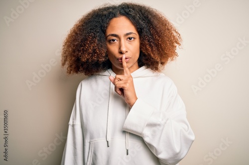 Young african american sportswoman with afro hair wearing sporty sweatshirt asking to be quiet with finger on lips. Silence and secret concept.