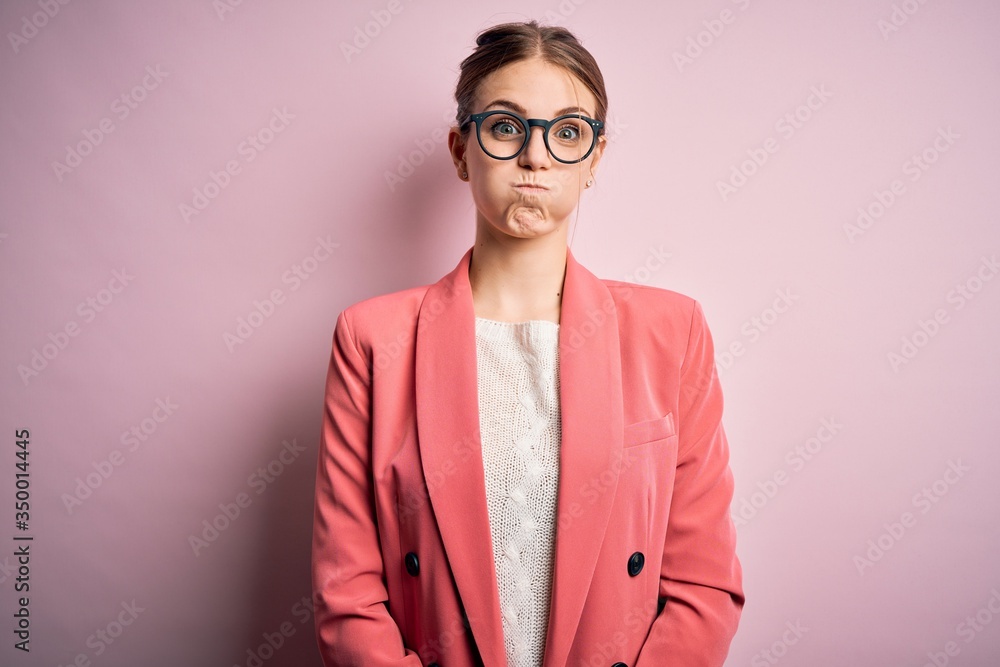 Young beautiful redhead woman wearing jacket and glasses over isolated pink background puffing cheeks with funny face. Mouth inflated with air, crazy expression.