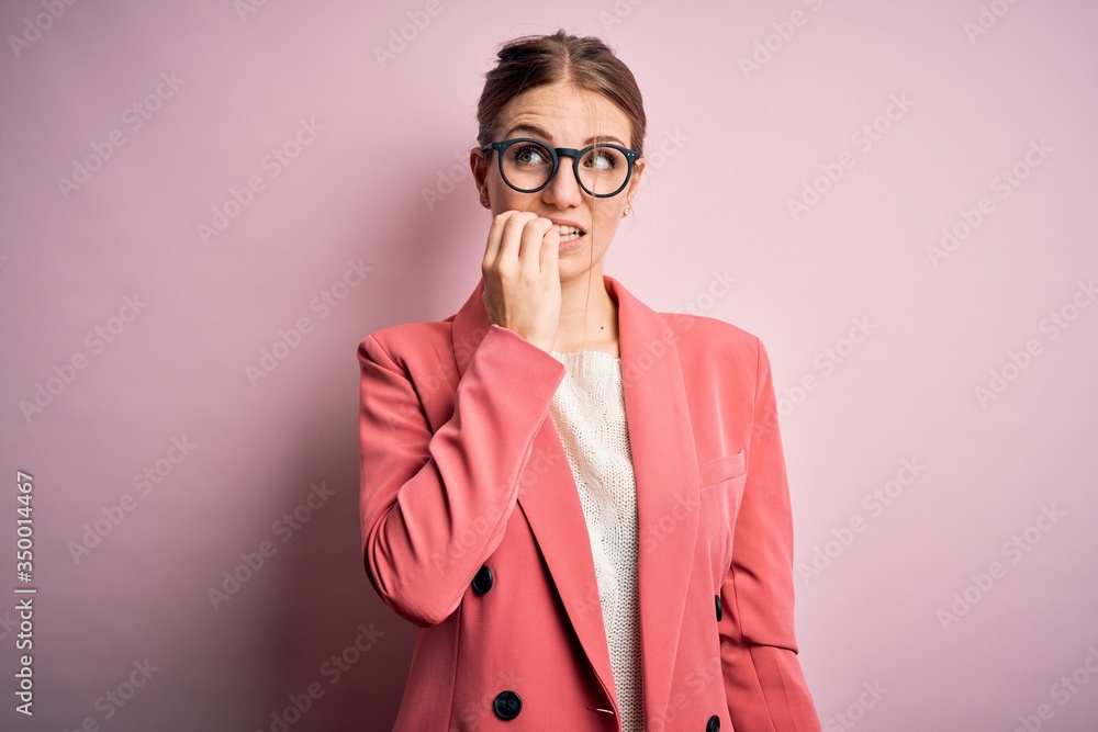 Young beautiful redhead woman wearing jacket and glasses over isolated pink background looking stressed and nervous with hands on mouth biting nails. Anxiety problem.