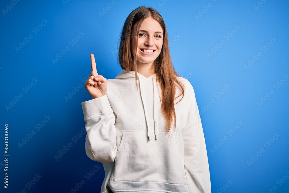Young beautiful redhead sporty woman wearing sweatshirt over isolated blue background showing and pointing up with finger number one while smiling confident and happy. Young beautiful redhead sporty woman wearing sweatshirt over isolated blue background showing and pointing up with finger number one while smiling confident and happy.