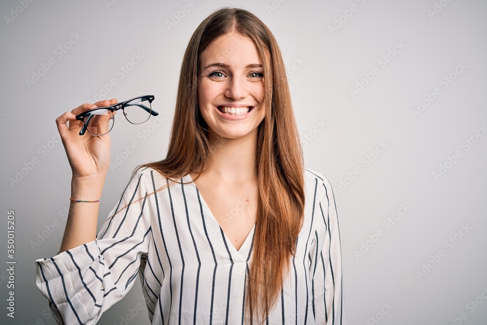 Young beautiful redhead optical woman holding glasses over isolated white background with a happy face standing and smiling with a confident smile showing teeth Young beautiful redhead optical woman holding glasses over isolated white background with a happy face standing and smiling with a confident smile showing teeth