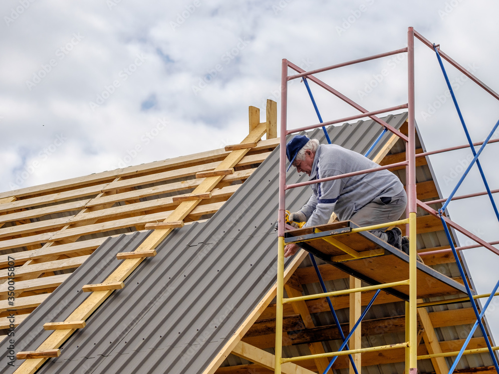 Naklejka premium Senior gray-haired Construction man using a screwdriver, fastens a roofing sheet to wooden rafters on the roof of a country house under construction. Physical activity of the seniors.
