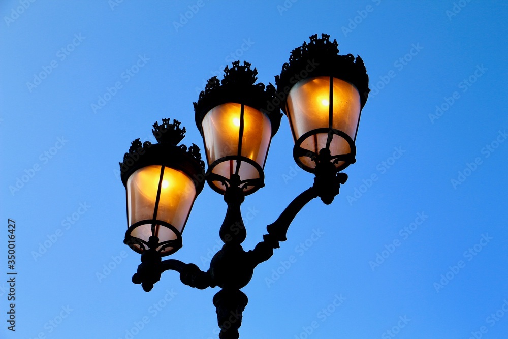 Surprising photo taken from below of a black antique wrought iron street light with 3 yellow lights lit in the daytime and with a beautiful blue sky without clouds in the background. Surprising photo taken from below of a black antique wrought iron street light with 3 yellow lights lit in the daytime and with a beautiful blue sky without clouds in the background.