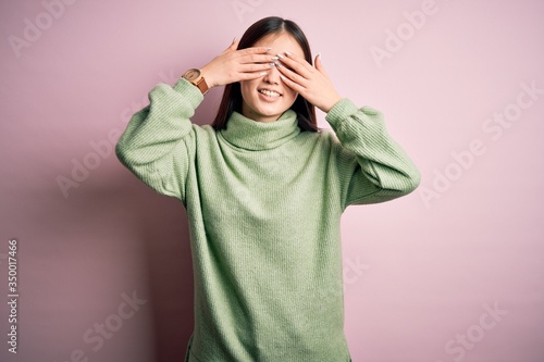 Young beautiful asian woman wearing green winter sweater over pink solated background covering eyes with hands smiling cheerful and funny. Blind concept. © Krakenimages.com