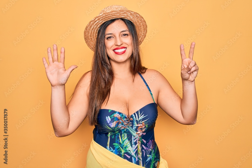 Young hispanic woman wearing summer hat and swimsuit over yellow background showing and pointing up with fingers number seven while smiling confident and happy. Young hispanic woman wearing summer hat and swimsuit over yellow background showing and pointing up with fingers number seven while smiling confident and happy.