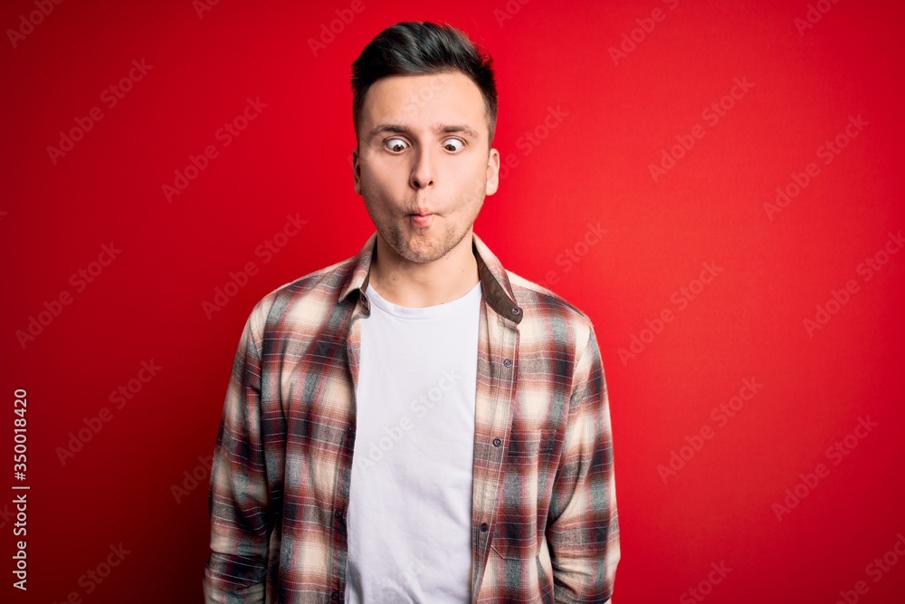 Obraz premium Young handsome caucasian man wearing casual modern shirt over red isolated background making fish face with lips, crazy and comical gesture. Funny expression.