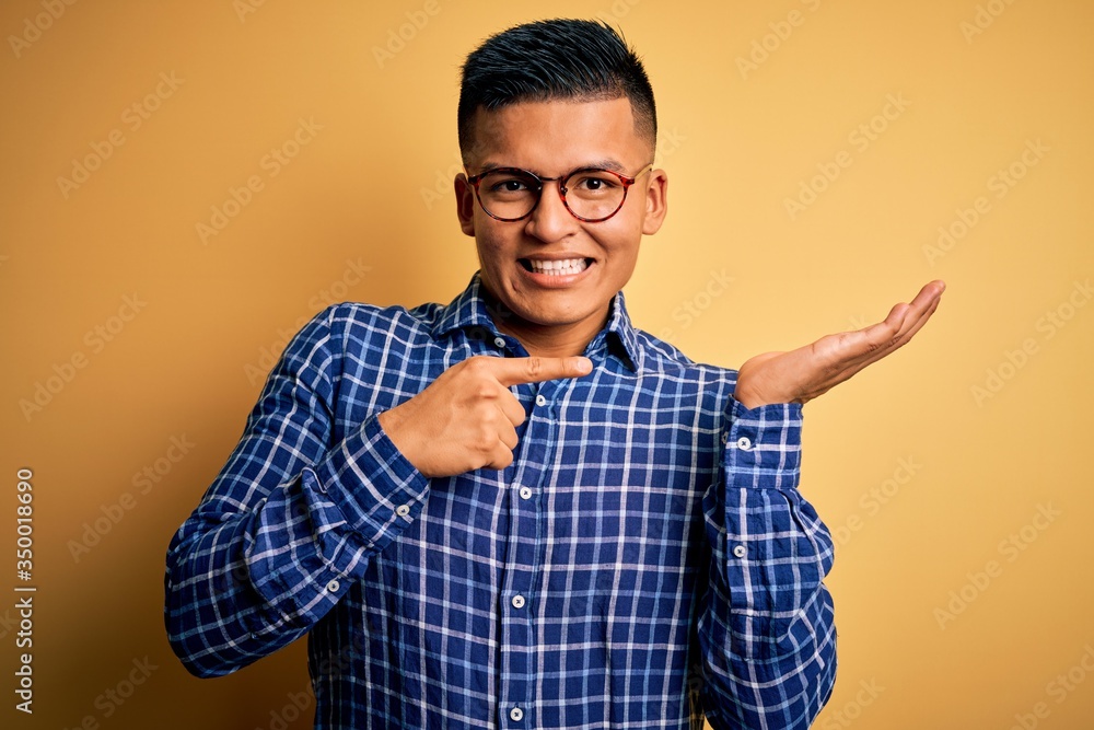 Young handsome latin man wearing casual shirt and glasses over yellow background amazed and smiling to the camera while presenting with hand and pointing with finger. Young handsome latin man wearing casual shirt and glasses over yellow background amazed and smiling to the camera while presenting with hand and pointing with finger.