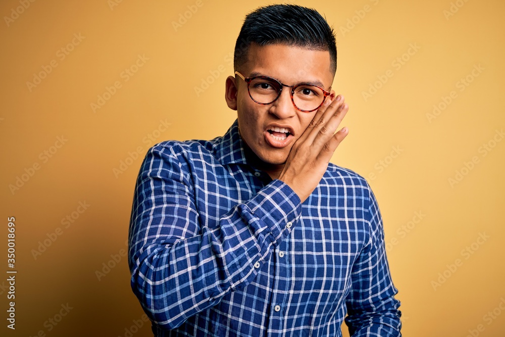 Young handsome latin man wearing casual shirt and glasses over yellow background hand on mouth telling secret rumor, whispering malicious talk conversation