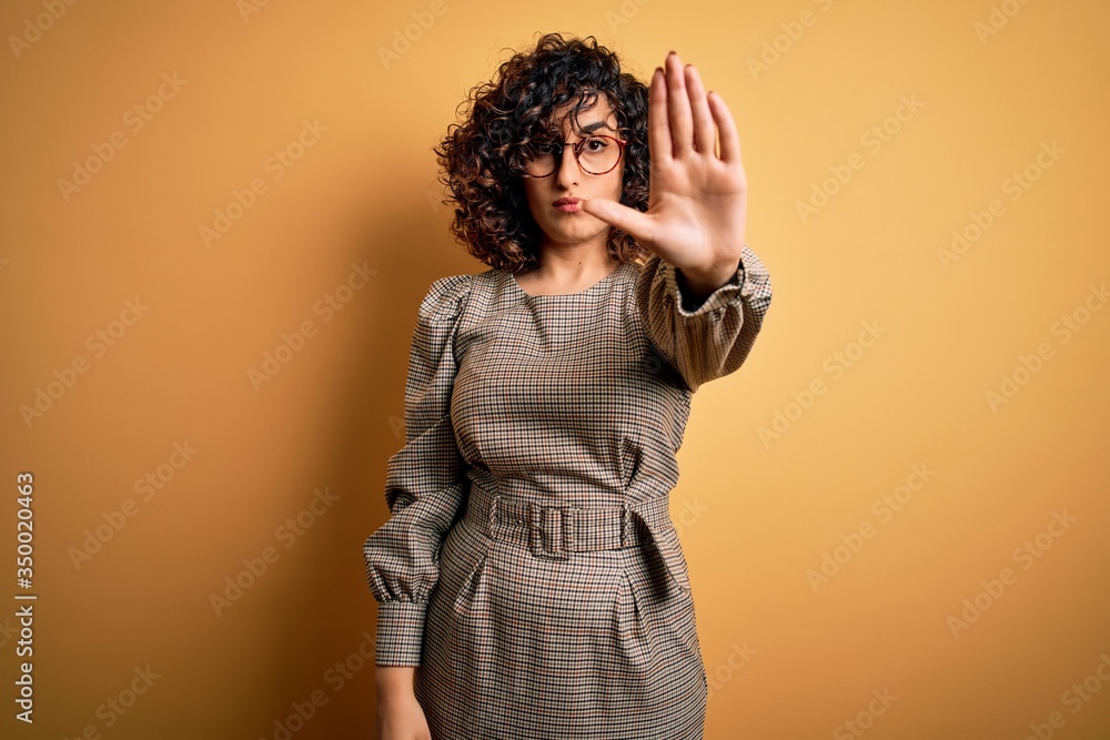 Beautiful arab business woman wearing dress and glasses standing over yellow background doing stop sing with palm of the hand. Warning expression with negative and serious gesture on the face. Beautiful arab business woman wearing dress and glasses standing over yellow background doing stop sing with palm of the hand. Warning expression with negative and serious gesture on the face.