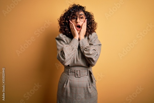 Beautiful arab business woman wearing dress and glasses standing over yellow background afraid and shocked, surprise and amazed expression with hands on face