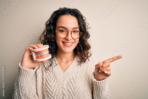 Young beautiful woman with curly hair holding plastic teeth with dental baces very happy pointing with hand and finger to the side photo