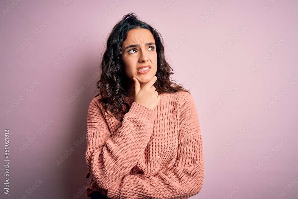 Young beautiful woman with curly hair wearing casual sweater over isolated pink background Thinking worried about a question, concerned and nervous with hand on chin Young beautiful woman with curly hair wearing casual sweater over isolated pink background Thinking worried about a question, concerned and nervous with hand on chin