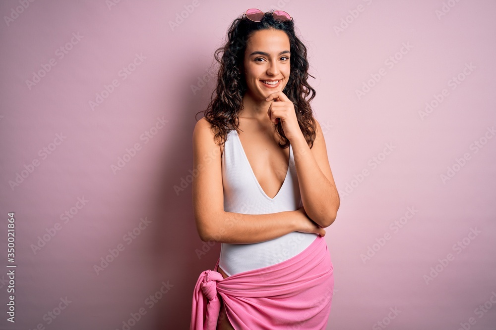 Obraz premium Beautiful woman with curly hair on vacation wearing white swimsuit over pink background looking confident at the camera smiling with crossed arms and hand raised on chin. Thinking positive.