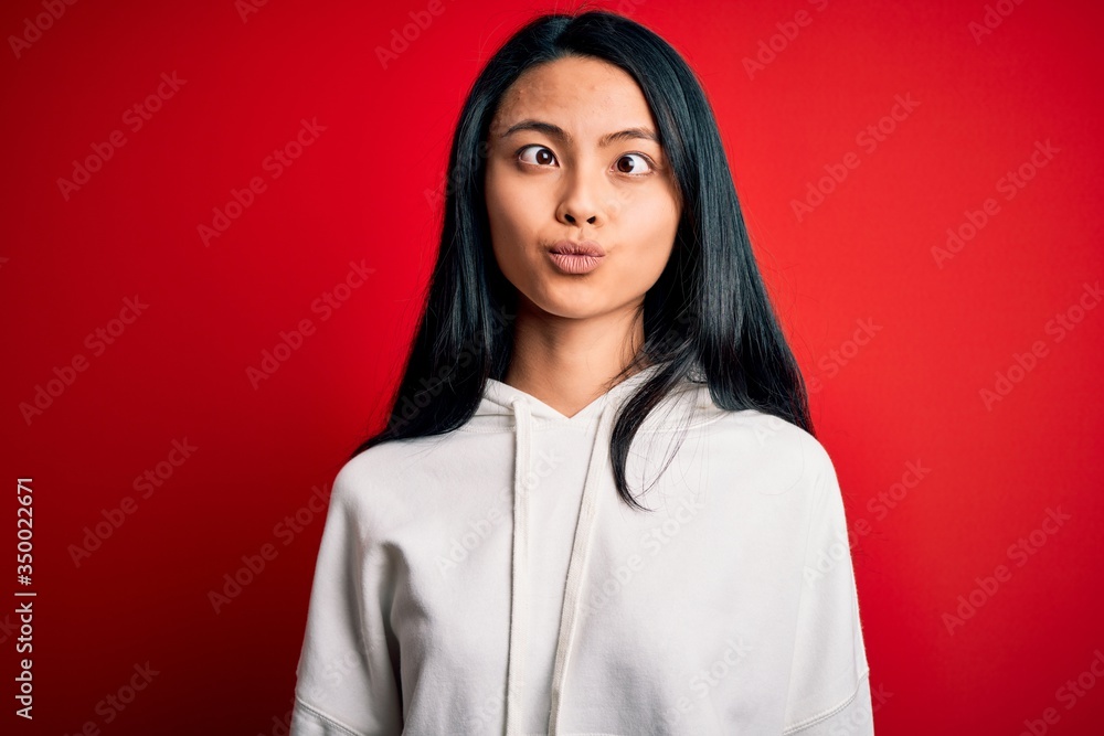 Young beautiful chinese sporty woman wearing sweatshirt over isolated red background making fish face with lips, crazy and comical gesture. Funny expression. Young beautiful chinese sporty woman wearing sweatshirt over isolated red background making fish face with lips, crazy and comical gesture. Funny expression.
