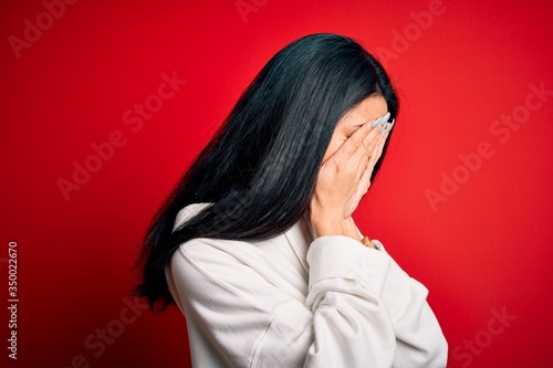 Young beautiful chinese sporty woman wearing sweatshirt over isolated red background with sad expression covering face with hands while crying. Depression concept.