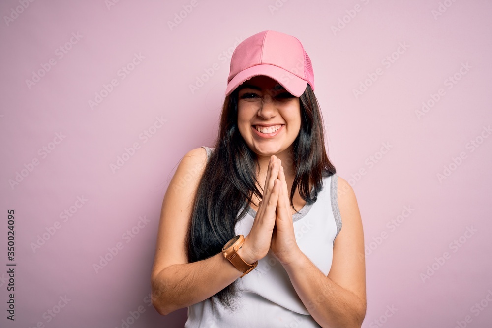 Young brunette woman wearing casual sport cap over pink background praying with hands together asking for forgiveness smiling confident. Young brunette woman wearing casual sport cap over pink background praying with hands together asking for forgiveness smiling confident.