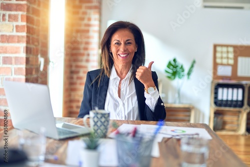 Middle age beautiful businesswoman working using laptop at the office doing happy thumbs up gesture with hand. Approving expression looking at the camera with showing success.